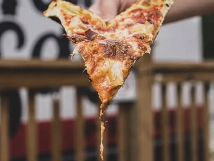 A slice of Mama Said pizza held up in front of the food truck